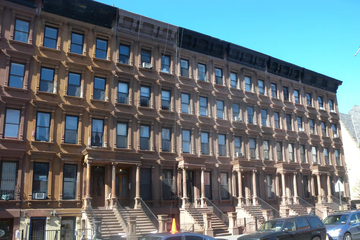 A long four-storey run of brownstone rowhouses with cornices and stoops on Lenox Avenue under a blue sky