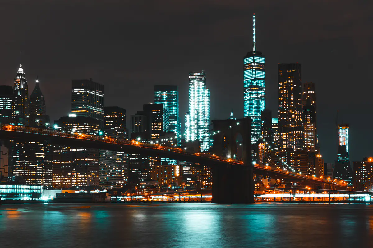The Brooklyn Bridge lit at night, the Lower Manhattan skyline and One World Trade Center glowing behind it and reflected in the river